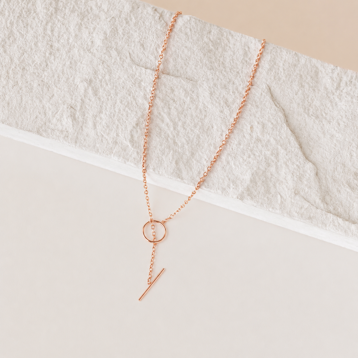 Rose gold necklace with circular and bar pendant on a textured white surface