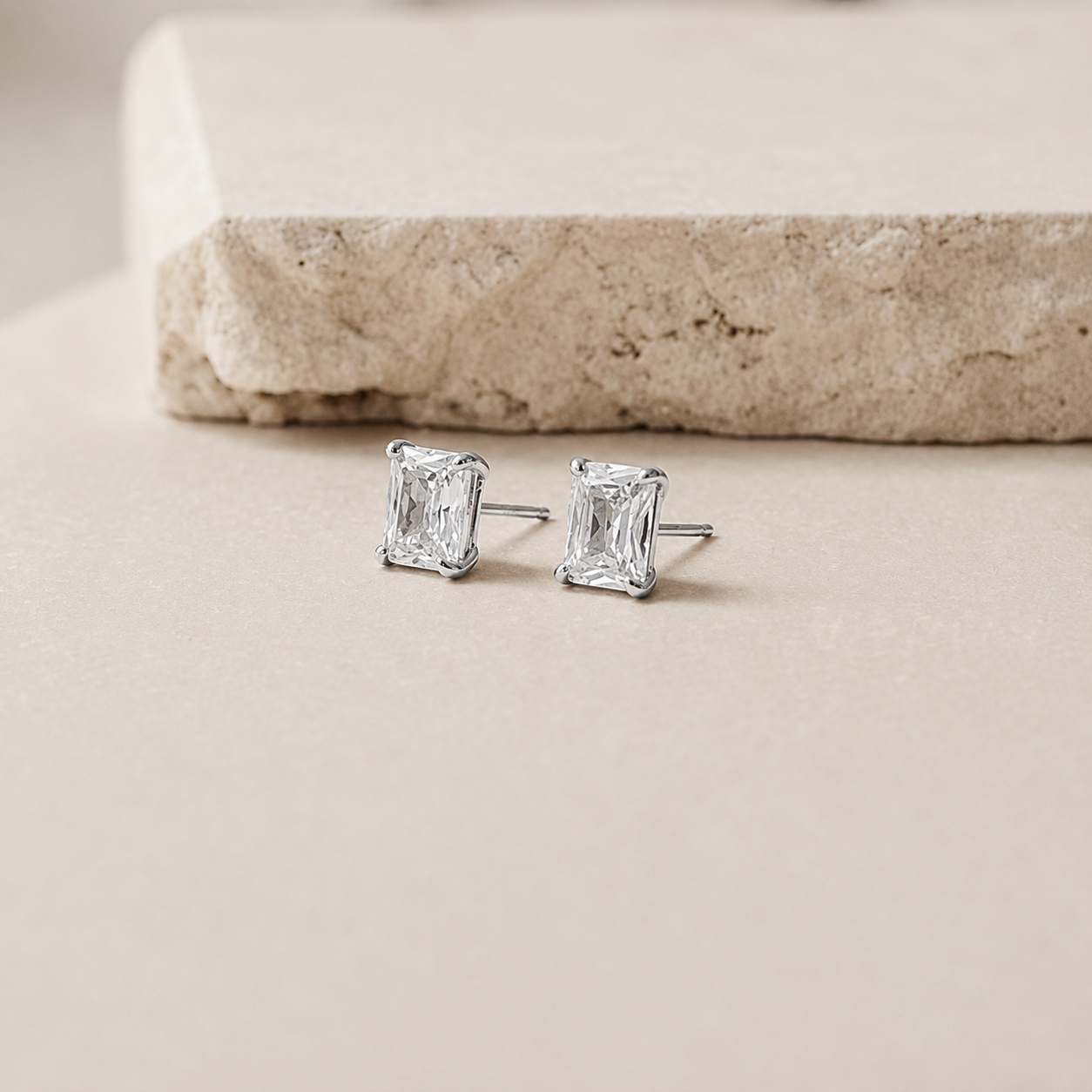 A pair of silver stud earrings with rectangular emerald-cut clear gemstones, resting on a light beige surface with a textured stone prop in the background.