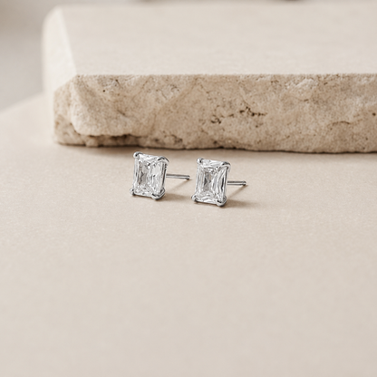 A pair of silver stud earrings with rectangular emerald-cut clear gemstones, resting on a light beige surface with a textured stone prop in the background.
