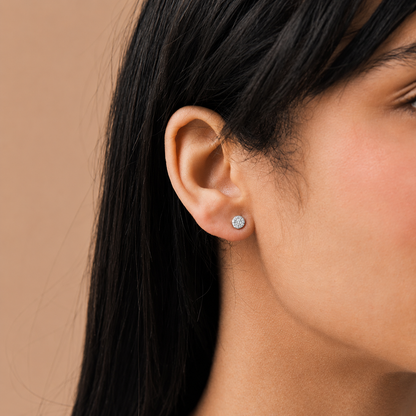 A close-up of a woman's ear wearing a small round stud earring with a clear gemstone in a silver-toned setting, against a warm beige background.