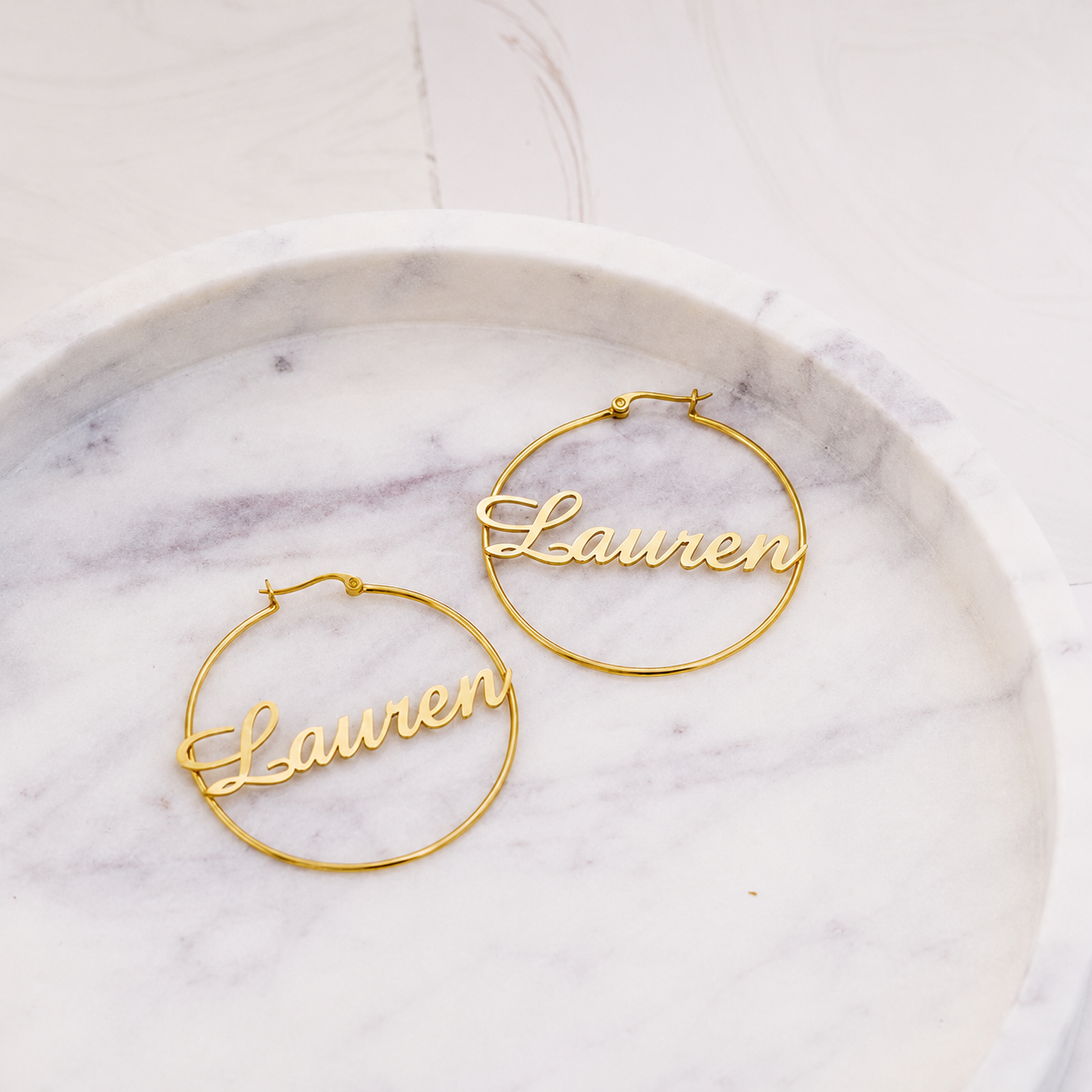 Gold hoop earrings with 'Lauren' engraving on a marble surface