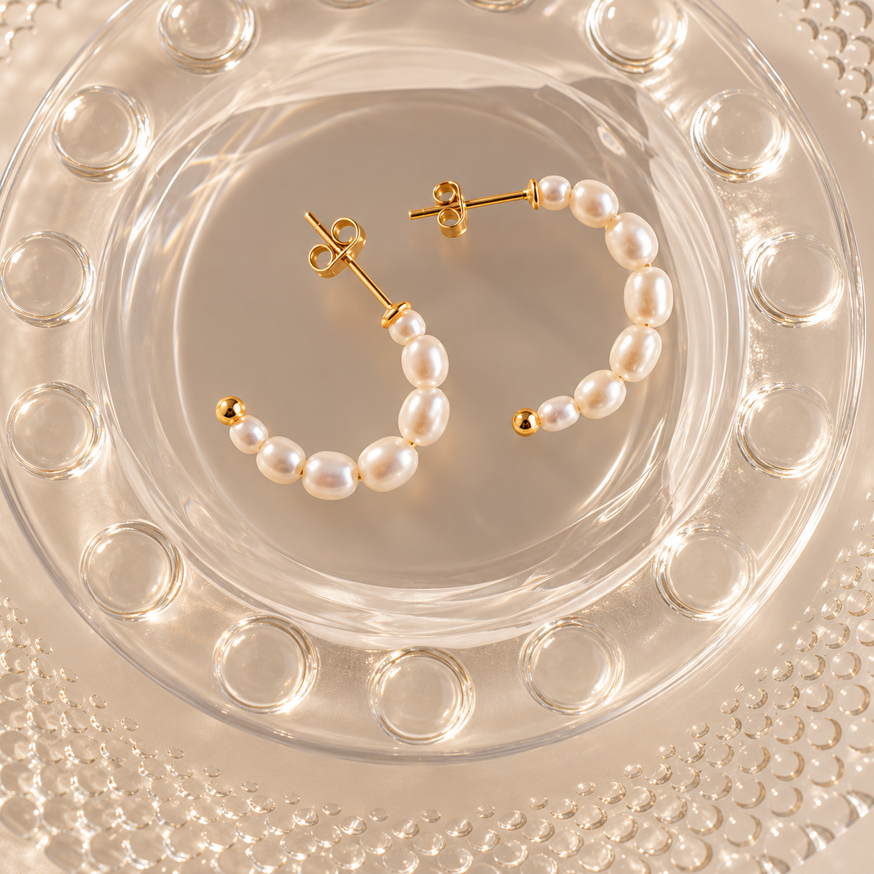 Gold hoop earrings with white pearl-like beads and small gold ball accents, displayed on a clear glass dish over a textured beige surface.