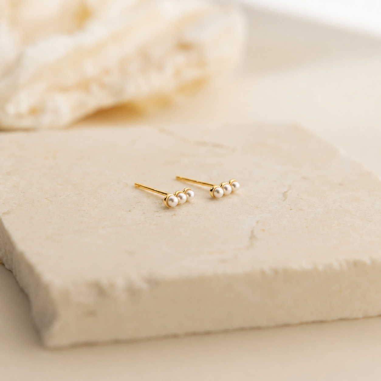 A pair of gold stud earrings with small white pearl-like beads, displayed on a light beige stone slab with a soft-focus background.
