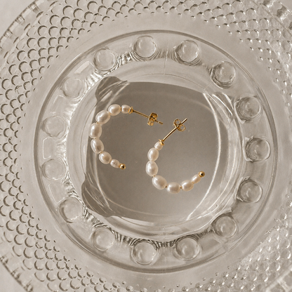 A pair of gold hoop earrings with small white pearl beads, displayed on a clear glass dish over a textured white surface.