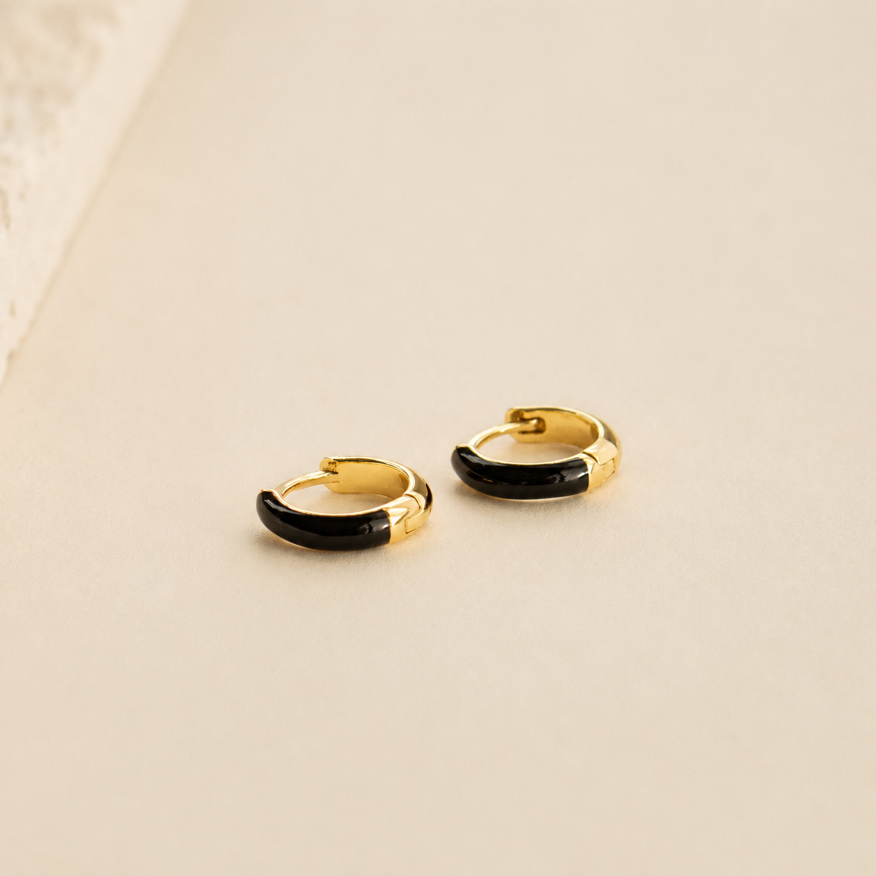 A pair of small gold huggie hoop earrings with a glossy black enamel inlay, displayed on a light beige surface.