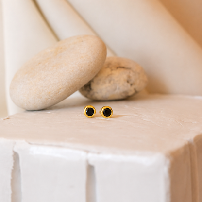 Pair of gold and black earrings on a white surface with stones in the background