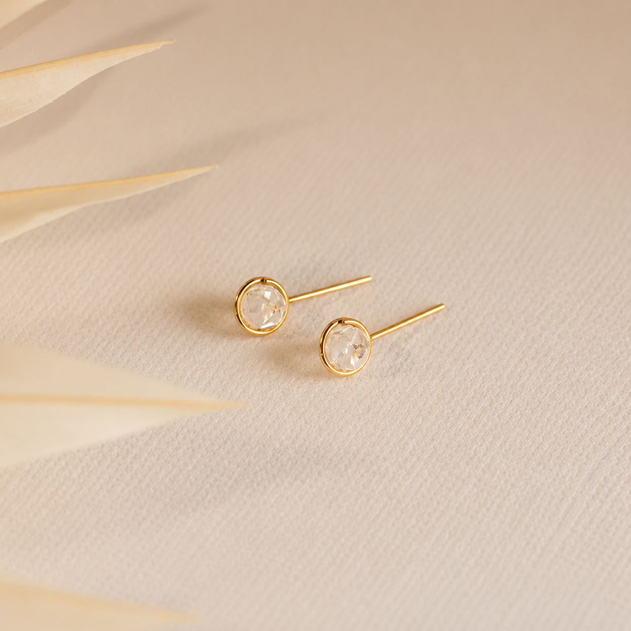 Gold stud earrings with clear gemstones on a beige fabric background