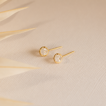 Gold stud earrings with clear gemstones on a beige fabric background