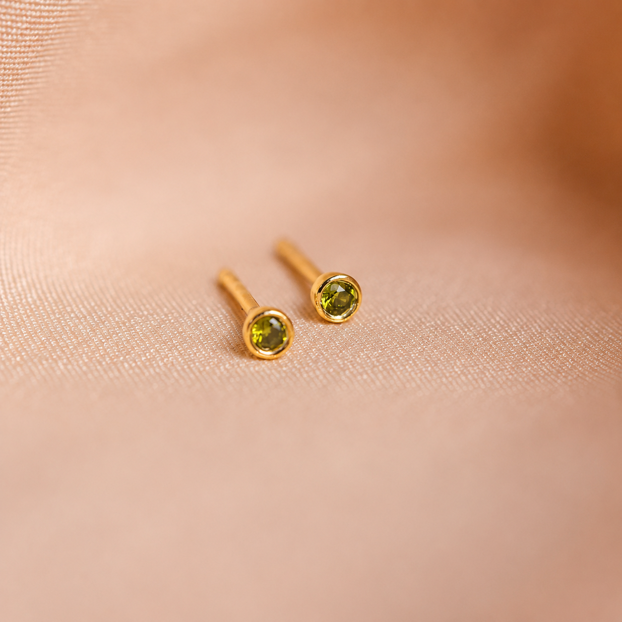 Gold stud earrings with green gemstones on a soft pink fabric background