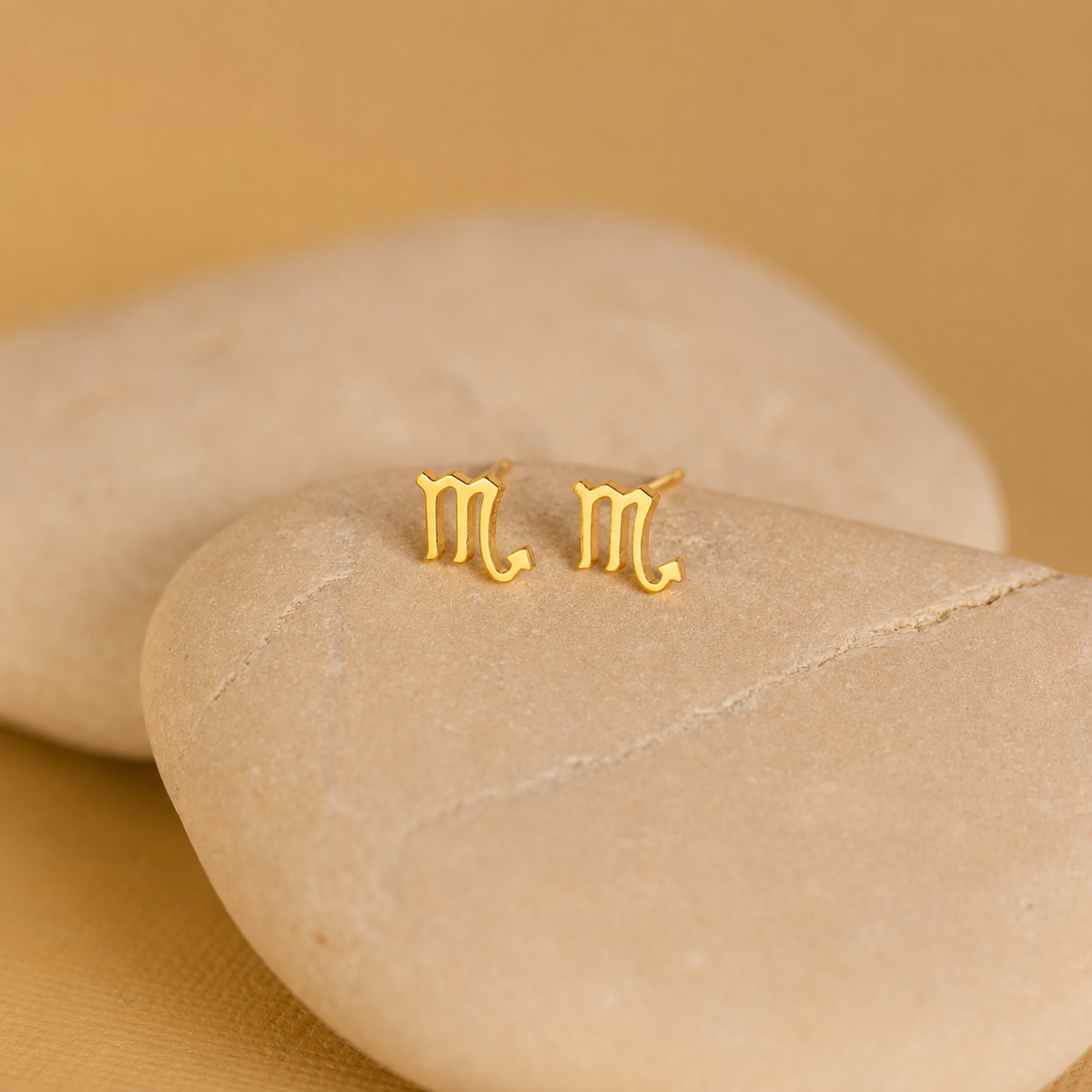 Gold 'M' earrings on a beige stone with a beige background