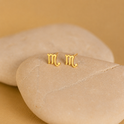 Gold 'M' earrings on a beige stone with a beige background