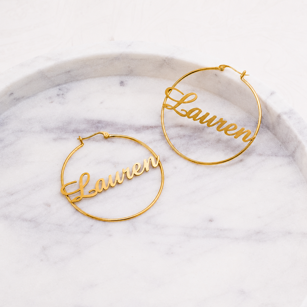 Gold hoop earrings with 'Lauren' engraved on a marble surface