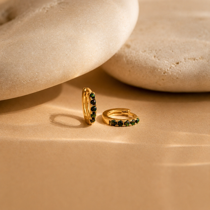 Gold hoop earrings with green gemstones on a beige stone surface