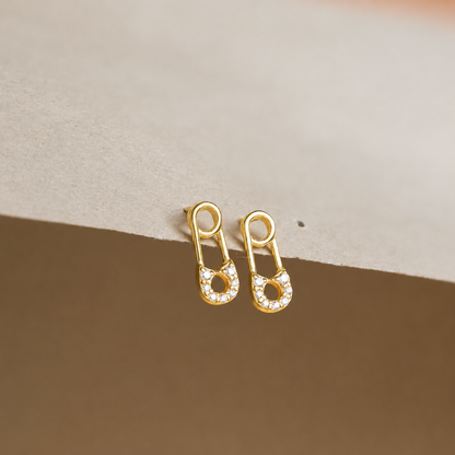 Gold safety pin earrings with clear stones on a beige background