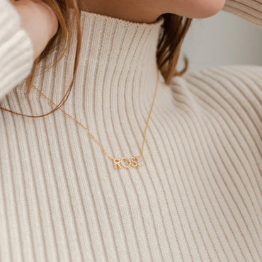 A person wearing a turtleneck sweater with a pave name necklace in rose gold tone.