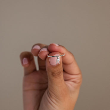 A hand holding a ring with a pave initial charm and a round-cut diamond birthstone charm on a plain background.