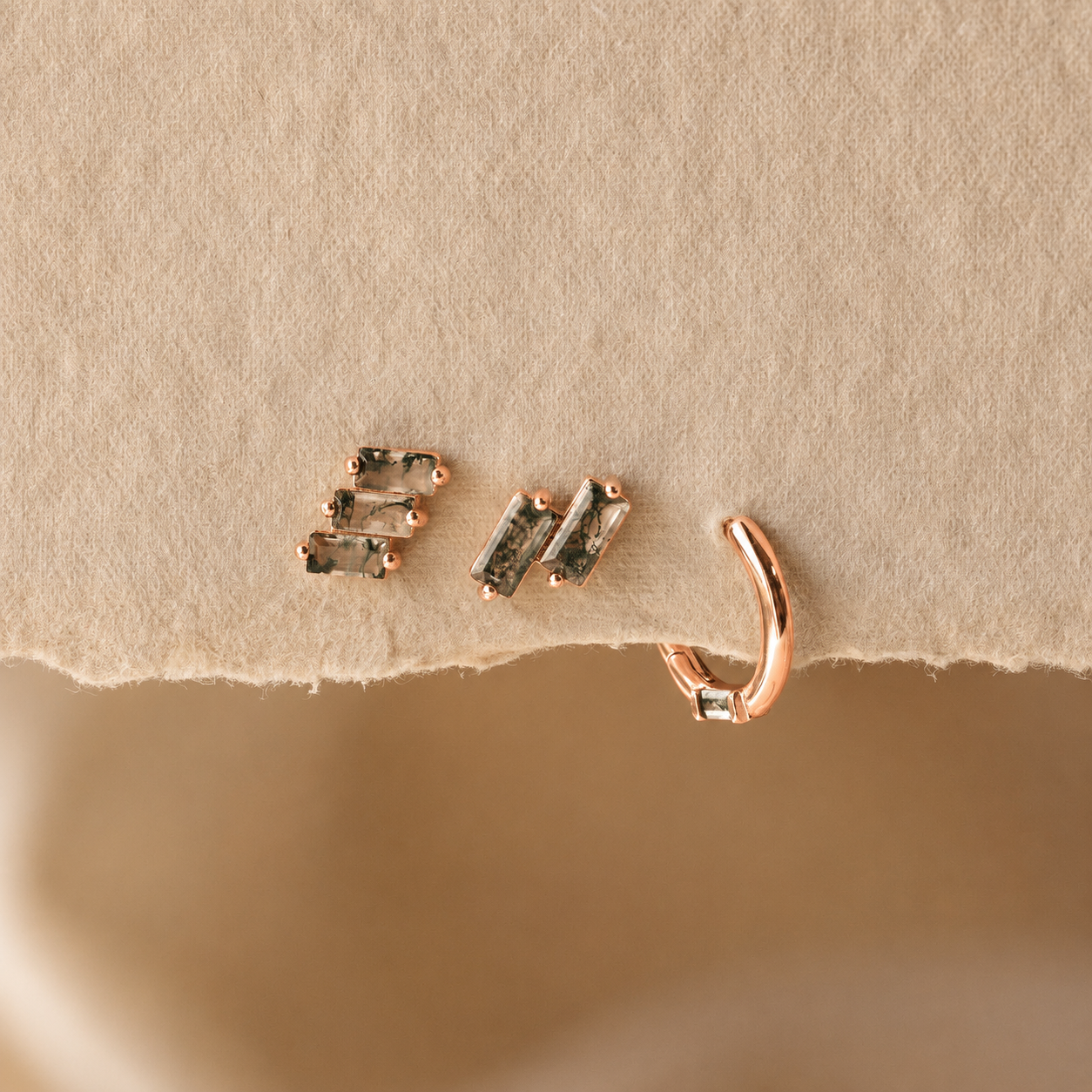 A close-up of rose gold jewelry displayed on a textured beige surface, featuring a small huggie hoop earring and a pair of stud earrings with dark rectangular stones.