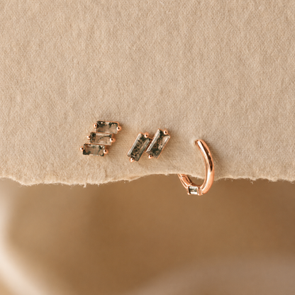 A close-up of rose gold jewelry displayed on a textured beige surface, featuring a small huggie hoop earring and a pair of stud earrings with dark rectangular stones.