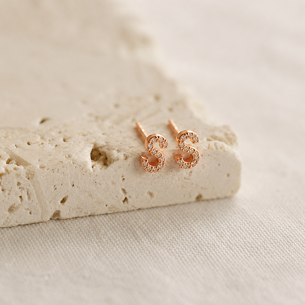 Rose gold letter 'S' earrings on a textured beige surface