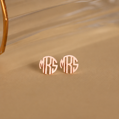 Rose gold 'MRS' earrings on a beige surface