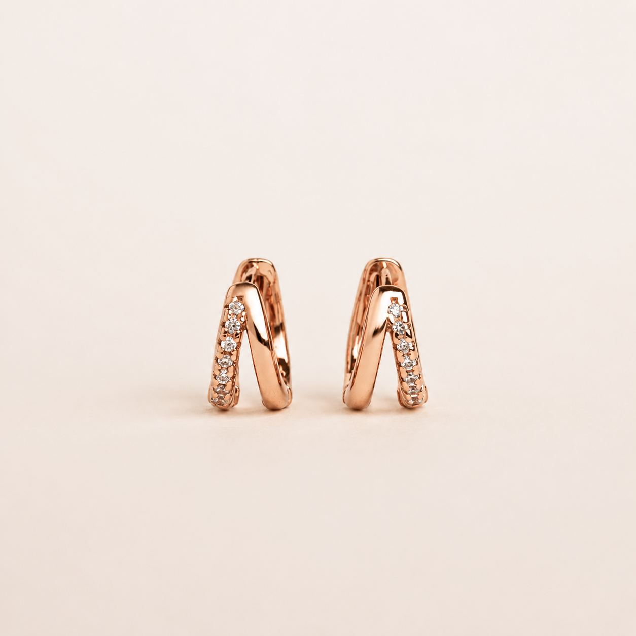 A pair of small rose gold huggie hoop earrings with pavé-set clear stones, shown on a clean, solid, light beige background.