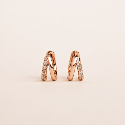 A pair of small rose gold huggie hoop earrings with pavé-set clear stones, shown on a clean, solid, light beige background.