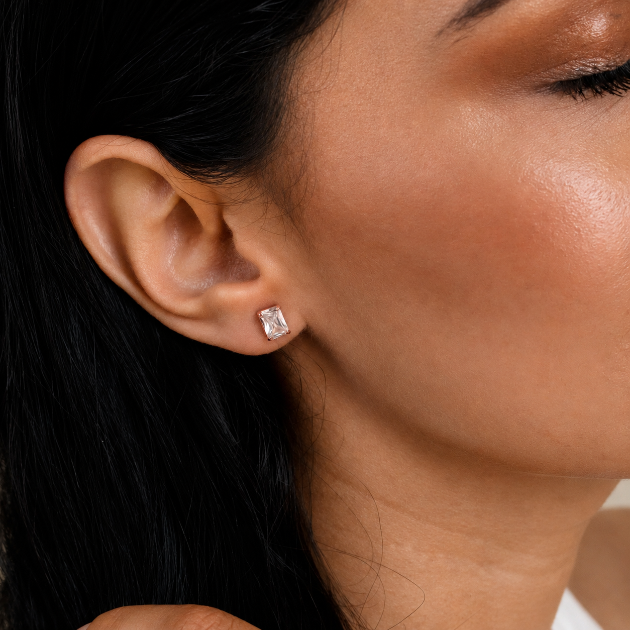 A close-up of a woman's ear wearing a small rectangular clear gemstone stud earring.