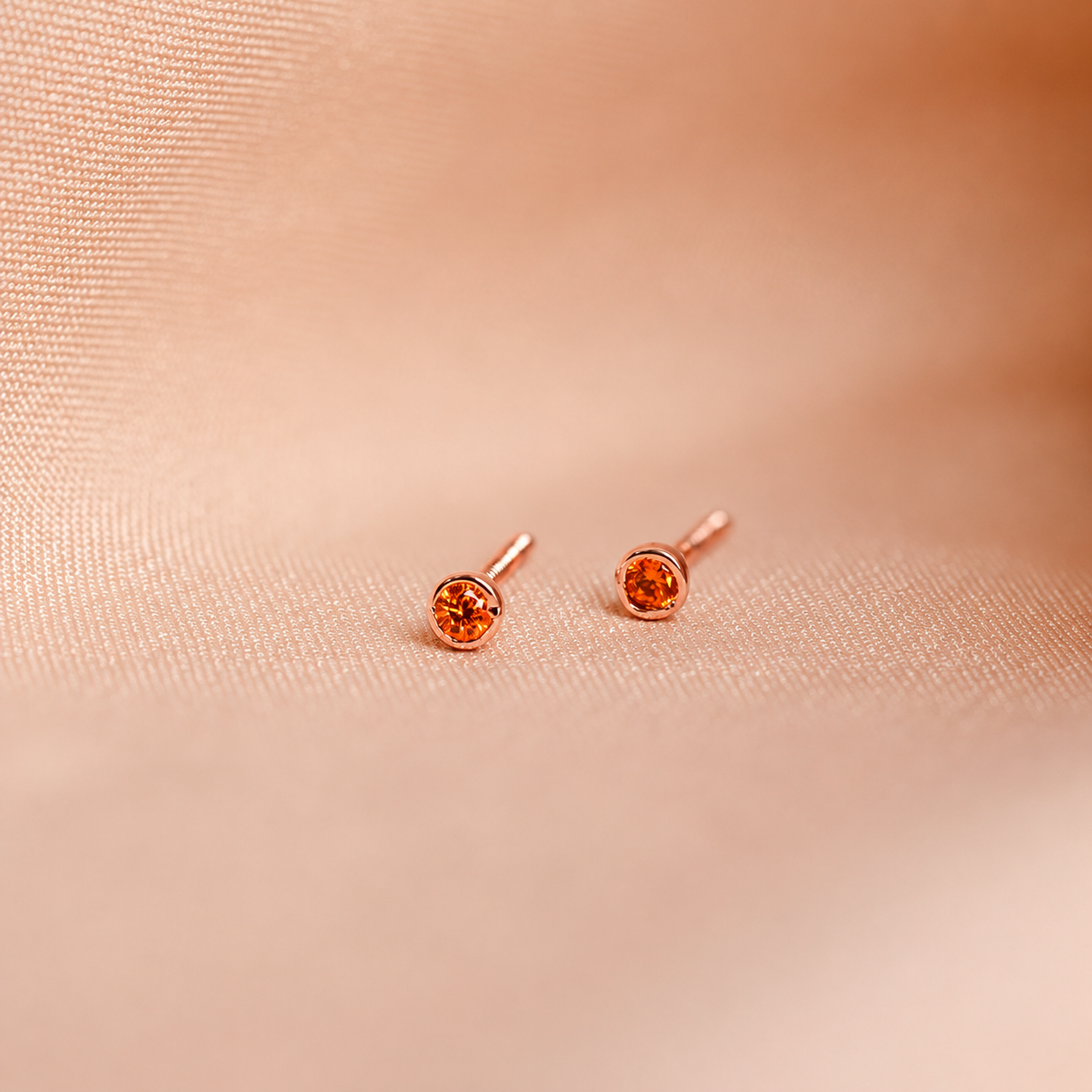 Rose gold earrings with orange gemstones on a soft pink fabric background