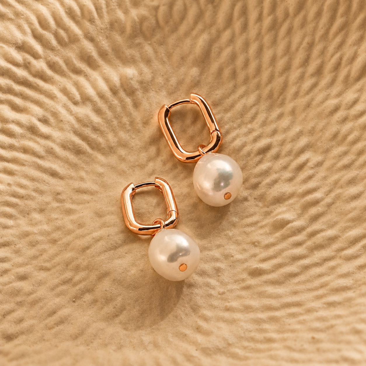 Pair of pearl earrings with rose gold hooks on a beige textured surface