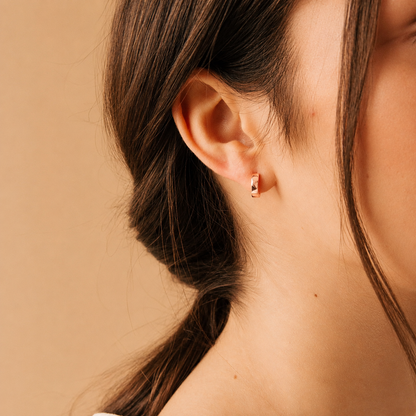 A close-up of a woman's ear wearing a small gold hoop earring against a warm beige background.