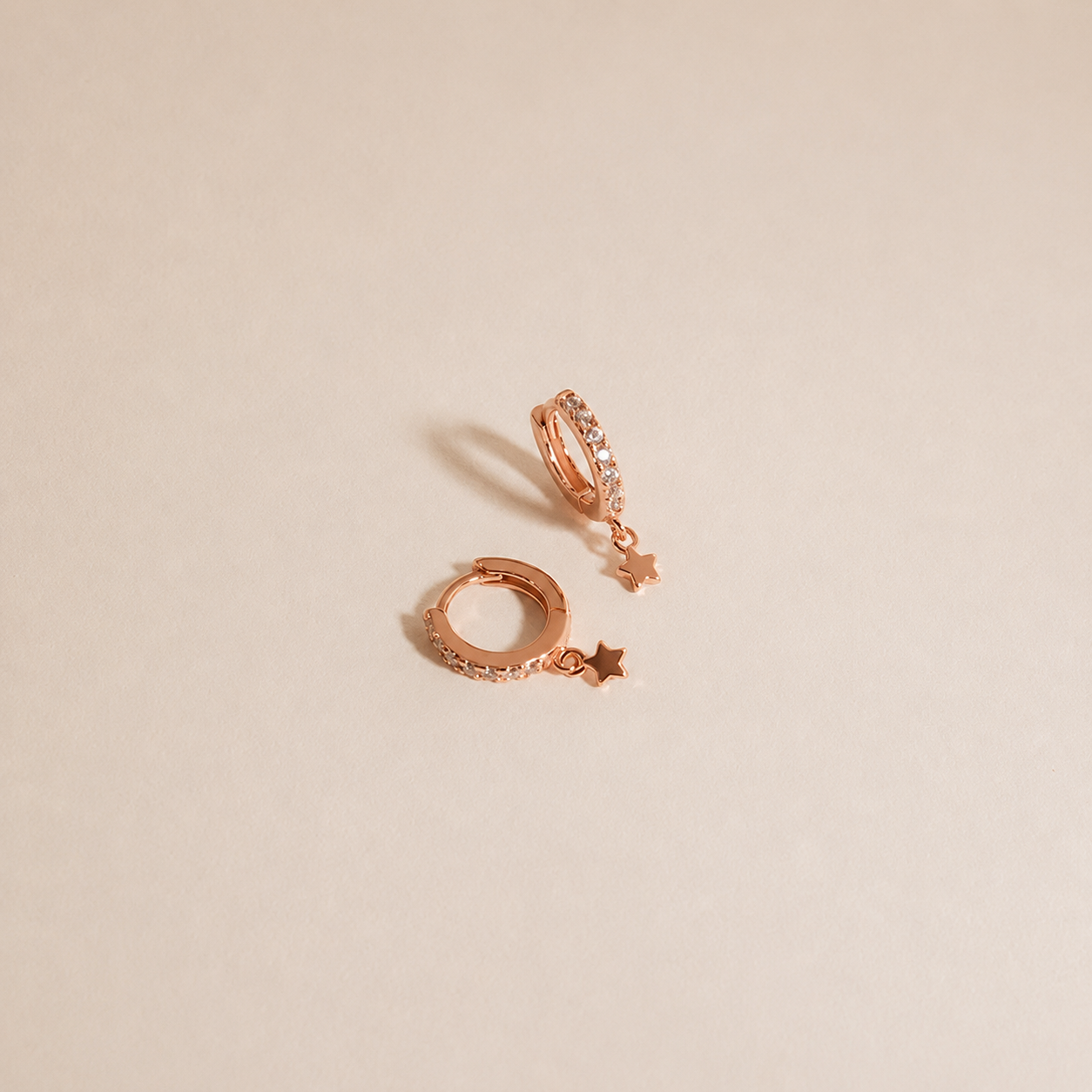 A pair of rose gold huggie hoop earrings with small dangling star charms, photographed on a clean, solid beige background.