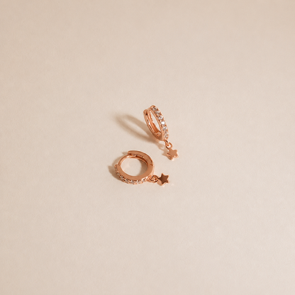 A pair of rose gold huggie hoop earrings with small dangling star charms, photographed on a clean, solid beige background.