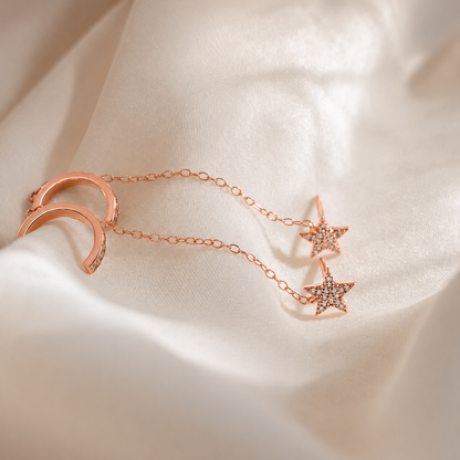 Rose gold jewelry set featuring a crescent moon and two star charms connected by a delicate chain, displayed on draped white satin fabric.