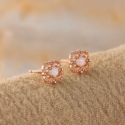 A pair of rose gold stud earrings with circular milgrain detailing and a central iridescent white opal gemstone, resting on a textured beige fabric surface.