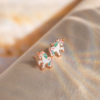 A pair of small unicorn stud earrings with white bodies, pink manes and tails, and mint green accents, set in rose gold metal and resting on a beige textured surface.