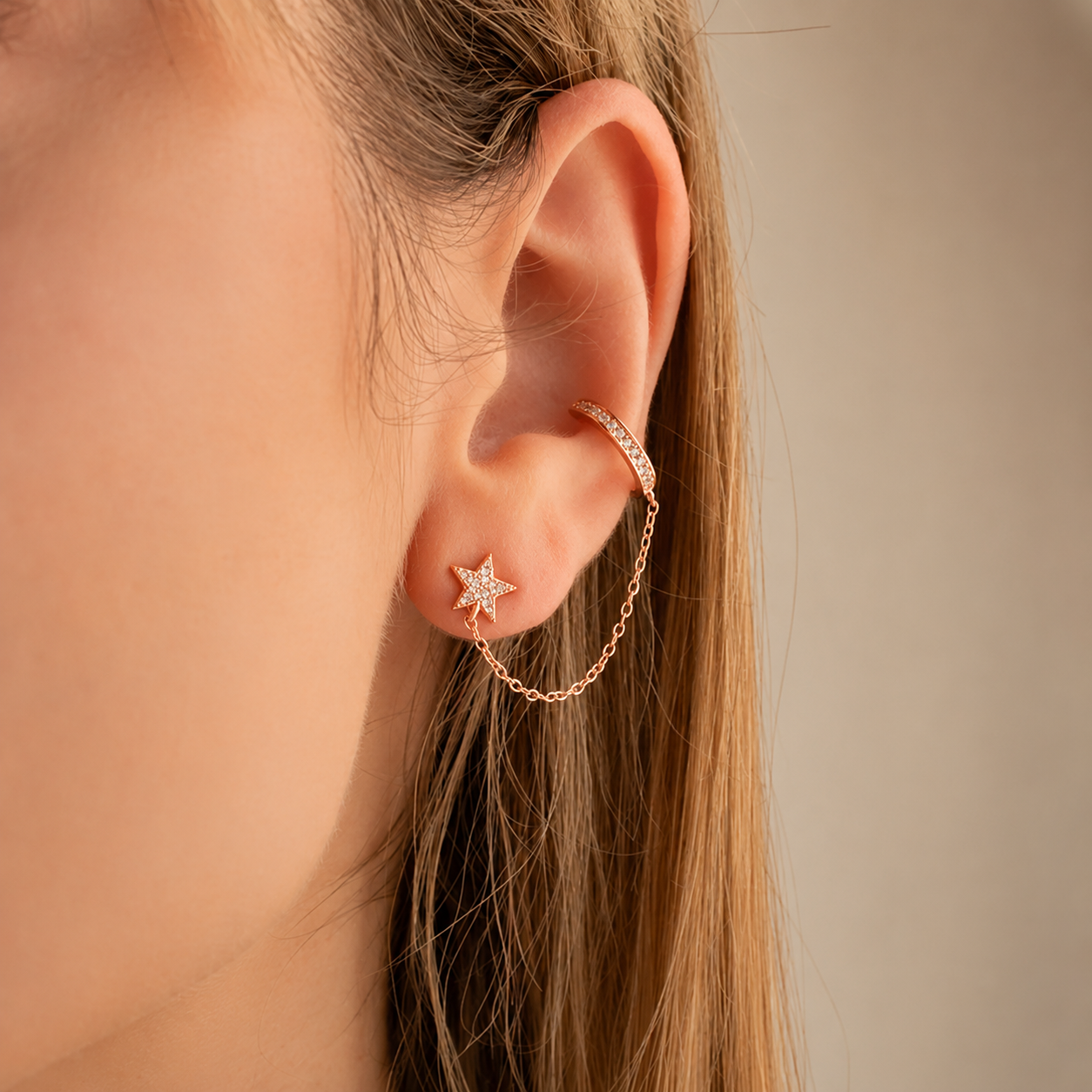 A close-up of a model's ear showcasing a rose gold ear cuff with a dangling chain and a small star-shaped stud earring.