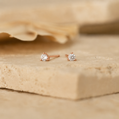 A pair of rose gold stud earrings with round clear gemstones, resting on a textured beige stone slab with a soft-focus background.