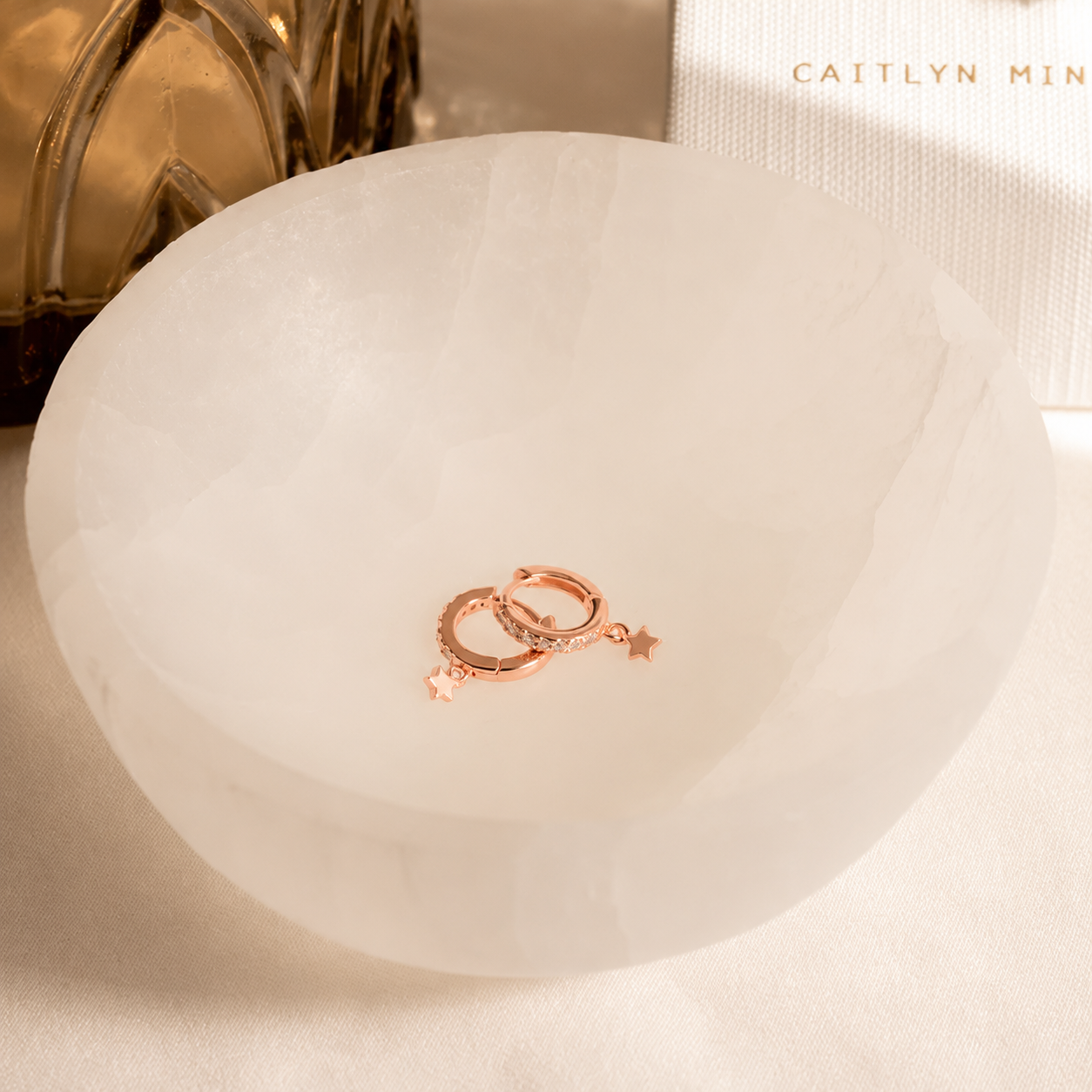 A pair of rose gold huggie hoop earrings with star charms, displayed inside a translucent white stone bowl on a neutral surface.