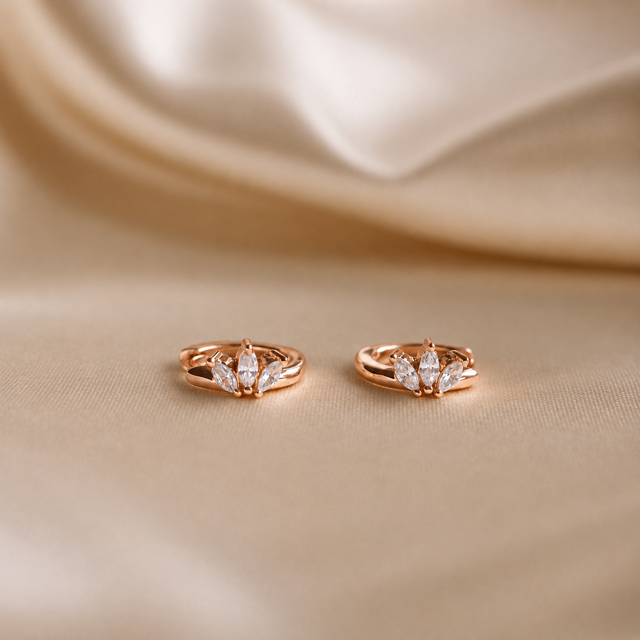 A pair of rose gold huggie hoop earrings with marquise-cut clear crystals, displayed on a draped beige satin fabric background.