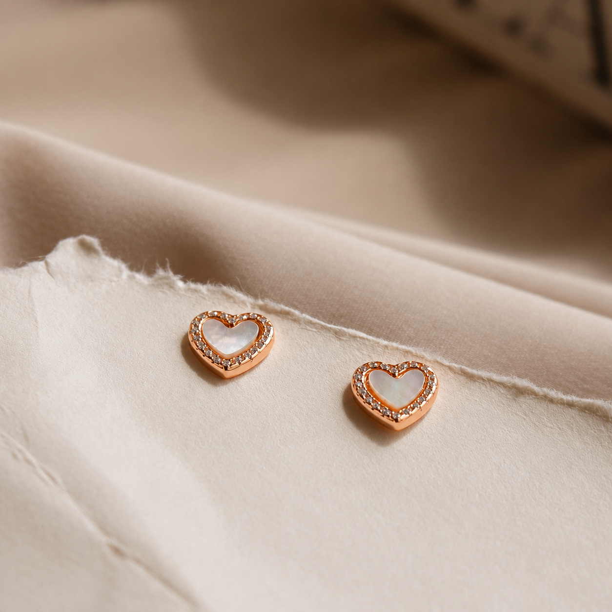 A pair of small heart-shaped stud earrings with rose gold borders and white inlay, resting on a beige fabric surface.
