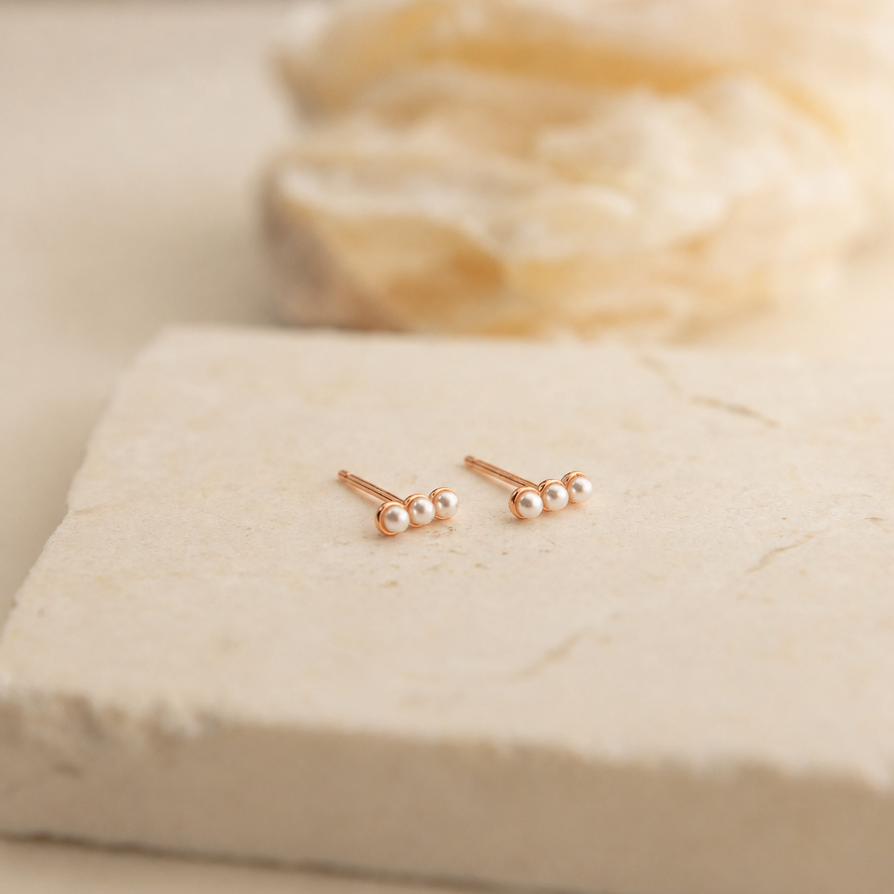 A pair of rose gold stud earrings with three small white pearls on each earring, displayed on a textured beige stone slab with a soft-focus background.