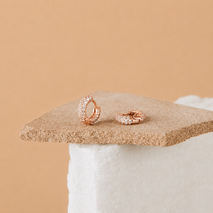 A pair of rose gold huggie hoop earrings paved with small clear stones, displayed on a textured tan stone slab over a white block against a solid tan background.