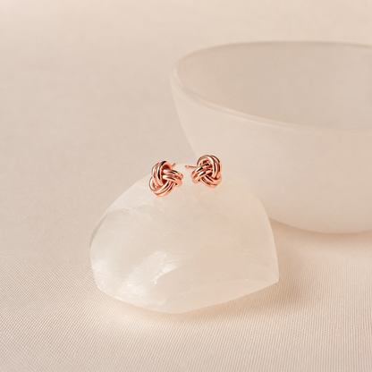 A pair of small knotted post earrings in polished rose gold, displayed on a white crystal cluster with a soft-focus white bowl in the background.