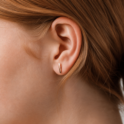 A close-up of a person's ear wearing a slim, vertical rose gold stud earring.