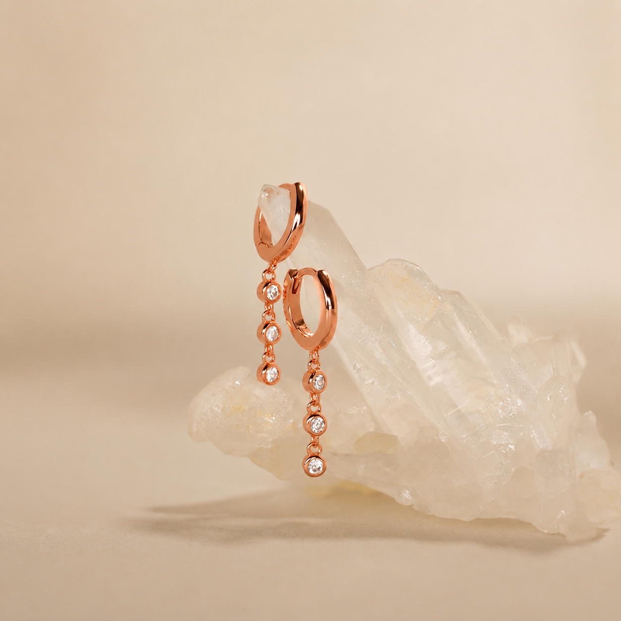 Rose gold hoop earrings with dangling crystals on a beige background