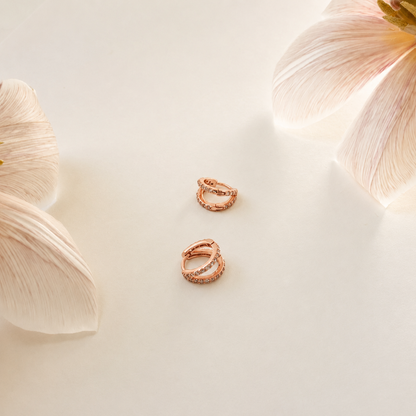 Two rose gold textured hoop earrings arranged on a light cream surface with soft white flower petals in the corners.
