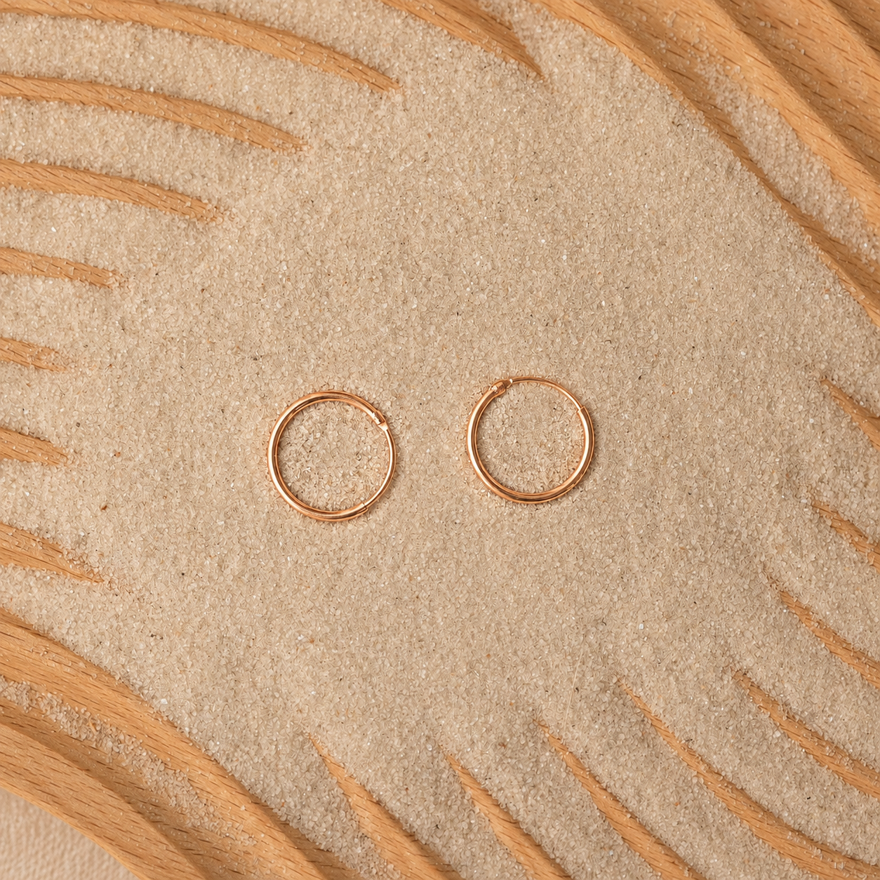 Two small thin hoop earrings in a warm metallic pink tone, placed on a textured beige surface with curved tan leaf-like lines around the edges.