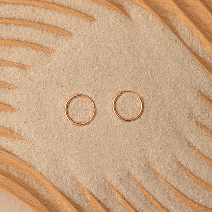Two small thin hoop earrings in a warm metallic pink tone, placed on a textured beige surface with curved tan leaf-like lines around the edges.
