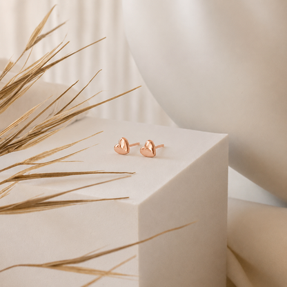 Rose gold heart-shaped earrings on a white pedestal with dried grasses and a white vase in the background.