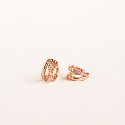 A pair of rose gold huggie hoop earrings with small clear pavé-set stones, displayed on a clean off-white background.
